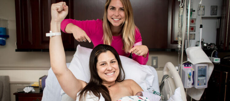 Położna wskazuje kobietę z noworodkiem. Kobieta podnosi rękę w geście radości i triumfu.